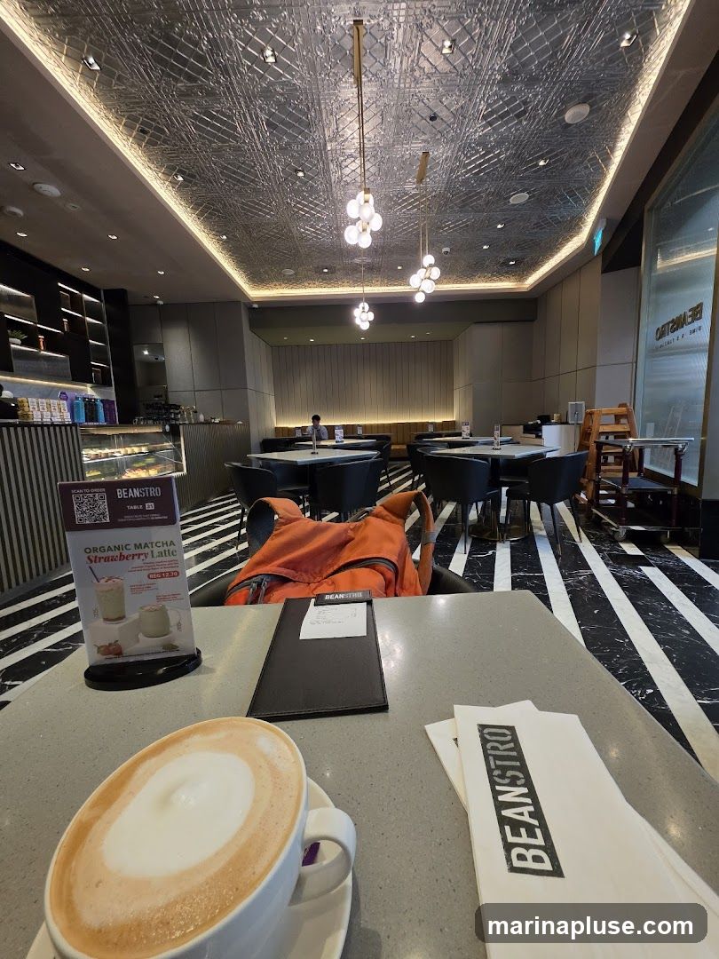 A view from a table in a modern cafe, with a latte in the foreground. The cafe has a distinctive ceiling with metallic textured panels and modern pendant lights. The floor has a black and white striped pattern. Tables and chairs are set up for customers, and a display case with pastries is visible in the background. A menu stand with a promotion for