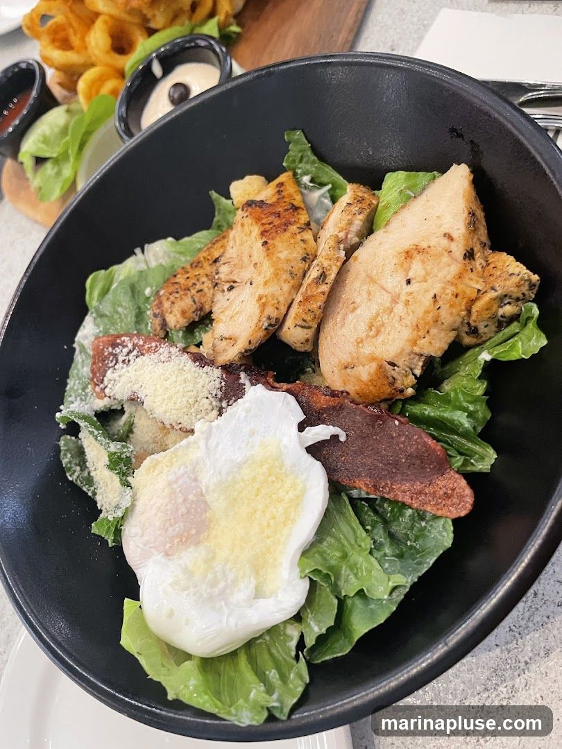 A close-up shot of a chicken Caesar salad in a black bowl. The salad contains grilled chicken slices, crispy bacon, a poached egg, grated cheese, and lettuce. In the background, there are curly fries and dipping sauces.