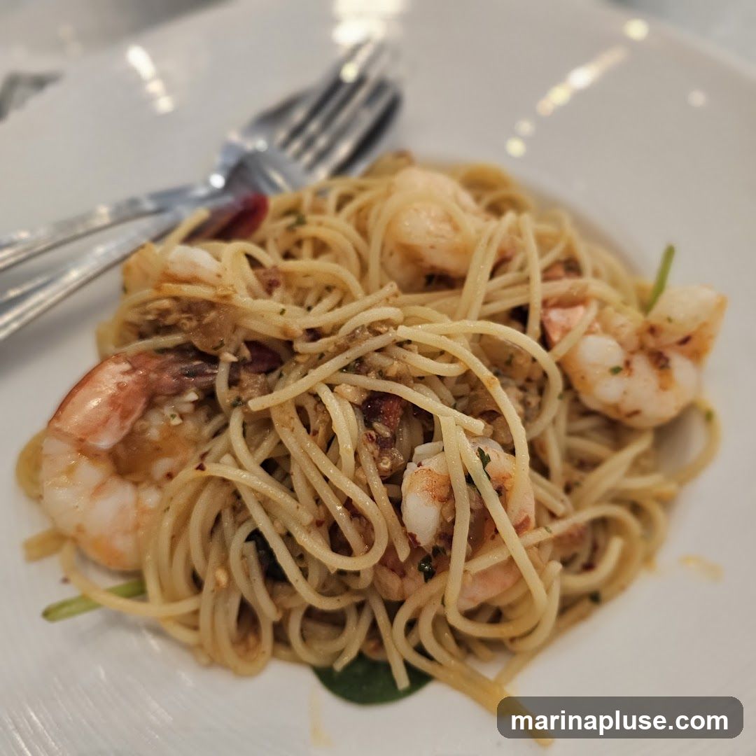 A close-up shot of spaghetti with shrimp, tossed in a light sauce with visible garlic and herbs. A fork and spoon are resting on the side of the white plate.
