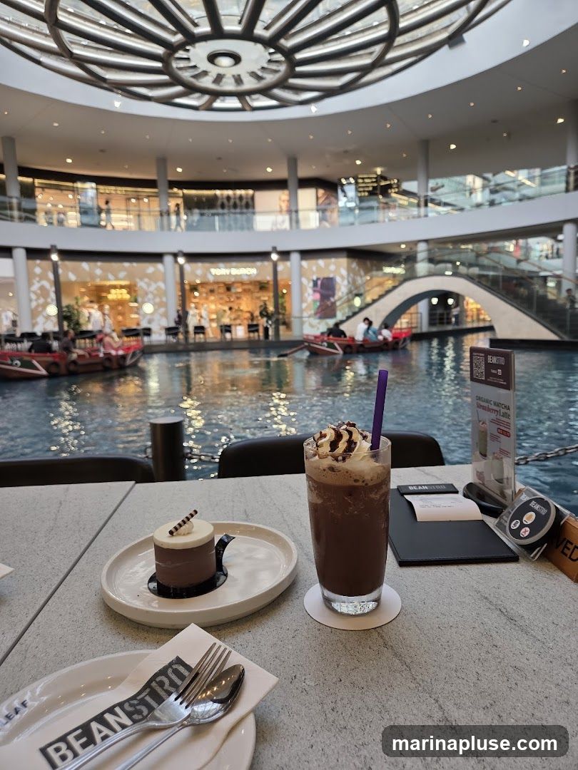 A table at a cafe with a small chocolate cake and a chocolate frappe. In the background, there is a canal with boats and shops in a large indoor atrium.