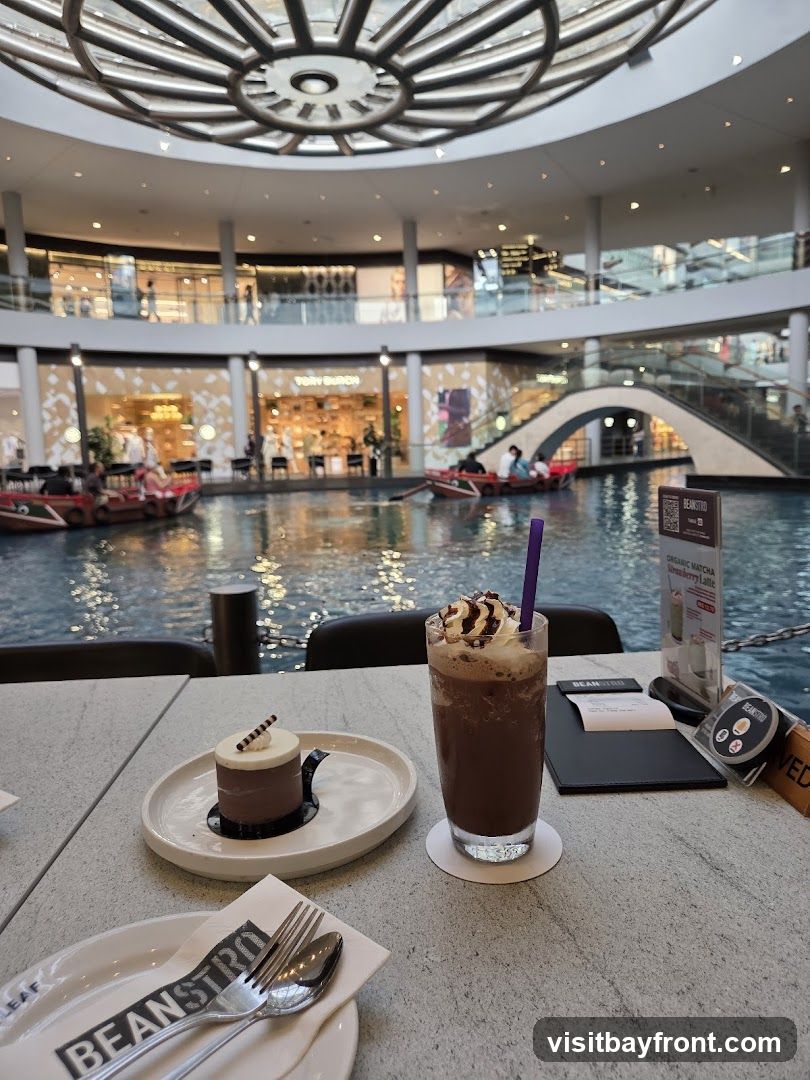A table at a cafe with a small chocolate cake and a chocolate frappe. In the background, there is a canal with boats and shops in a large indoor atrium.