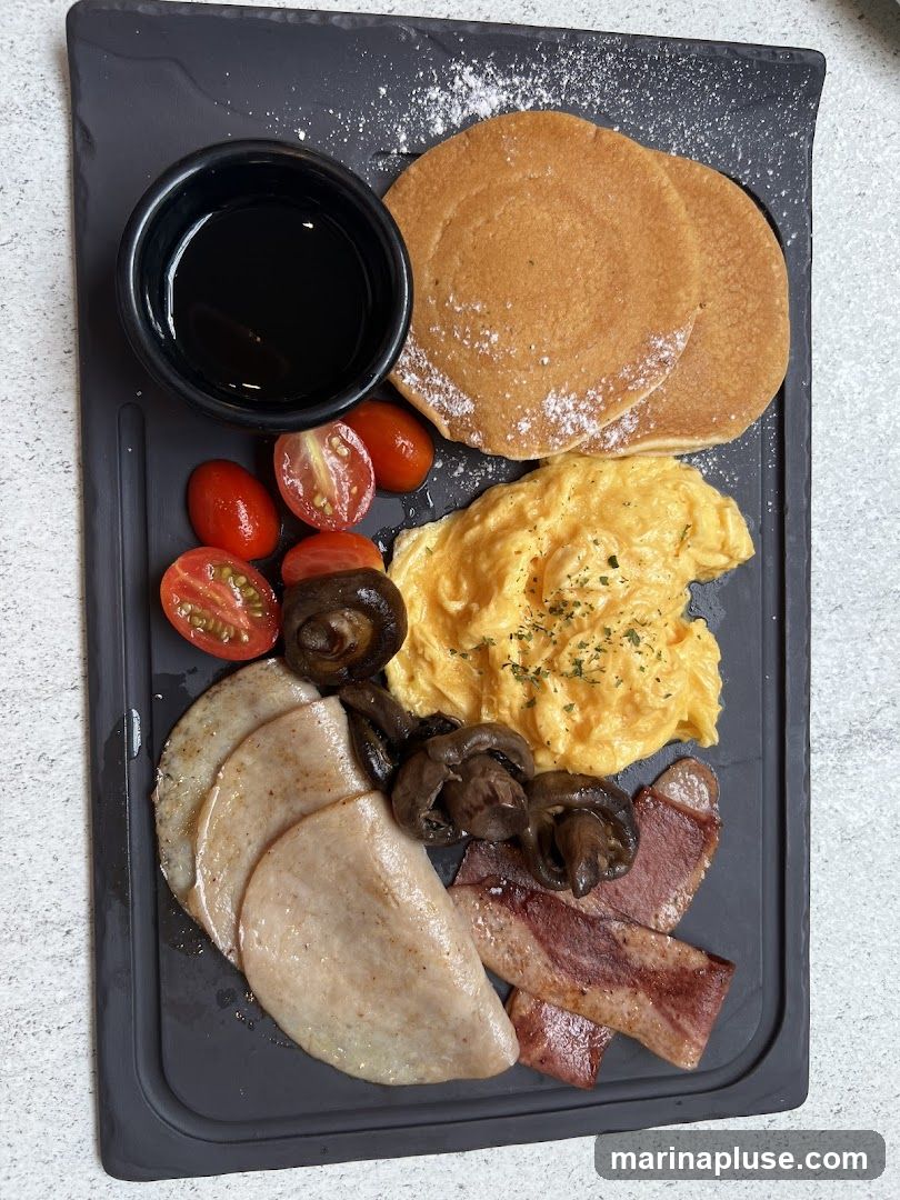 A breakfast platter served on a dark slate board. It includes two pancakes dusted with powdered sugar, a small bowl of syrup, scrambled eggs, cherry tomatoes, sautéed mushrooms, sliced turkey or ham, and bacon.