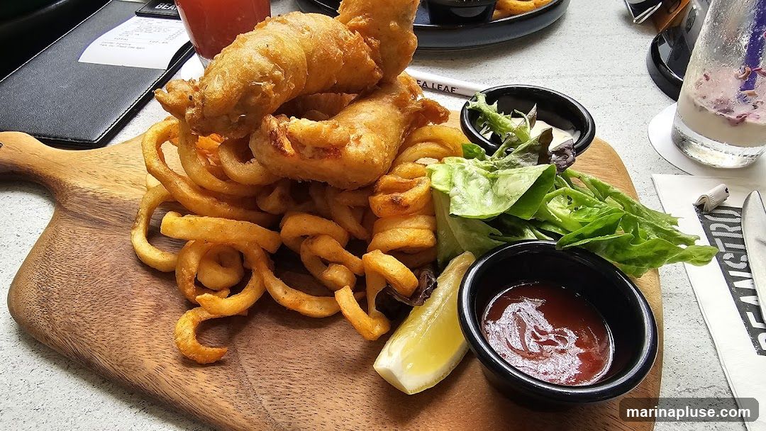 A close-up shot of a serving of fish and chips on a wooden board. The dish includes two pieces of battered fish, a large portion of curly fries, a small side salad, a wedge of lemon, and two dipping sauces (one appears to be ketchup, the other a white sauce). A drink and a menu are visible in the background.