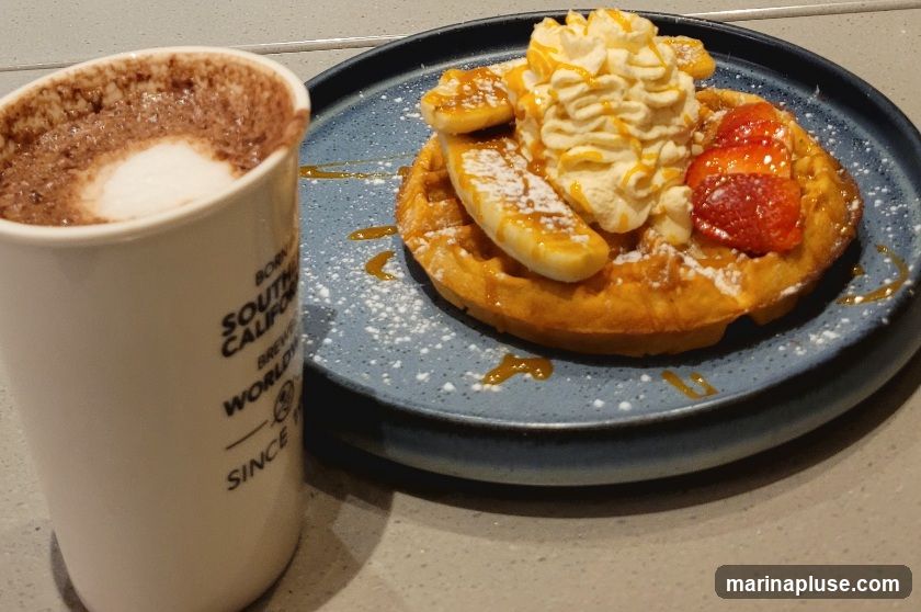 A close-up shot of a waffle topped with whipped cream, sliced bananas, and strawberries, drizzled with caramel sauce and dusted with powdered sugar. To the left, a white mug with a logo containing text like "BORN SOUTH CALIFORNIA BREW WORLDWIDE SINCE" holds a frothy beverage, likely hot chocolate or coffee.