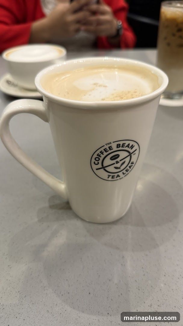 A white ceramic mug with the logo of "The Coffee Bean & Tea Leaf" is filled with a frothy coffee drink. In the background, another coffee drink in a white cup and a tall iced coffee drink are visible. A person wearing a red jacket is out of focus in the background.