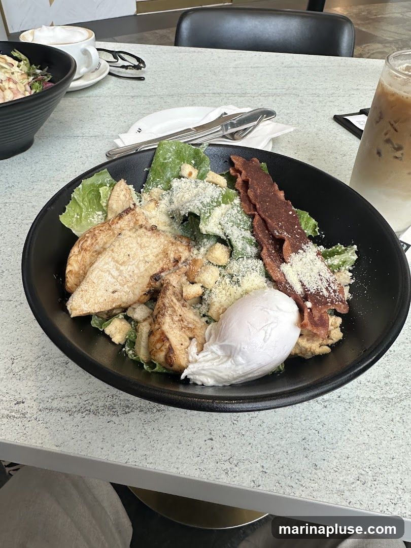 A close-up shot of a Caesar salad served in a black bowl. The salad contains lettuce, croutons, shredded cheese, grilled chicken slices, crispy bacon strips, and a poached egg. A glass of iced coffee and another salad bowl are visible in the background, along with a cup of coffee and eyeglasses on the table.