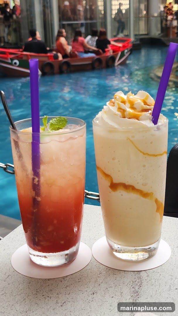 Two drinks are on a table with a body of water and a boat in the background. The drink on the left is a red, layered beverage with mint and a purple straw. The drink on the right is a creamy, light-colored beverage with whipped cream, caramel drizzle, and a purple straw.