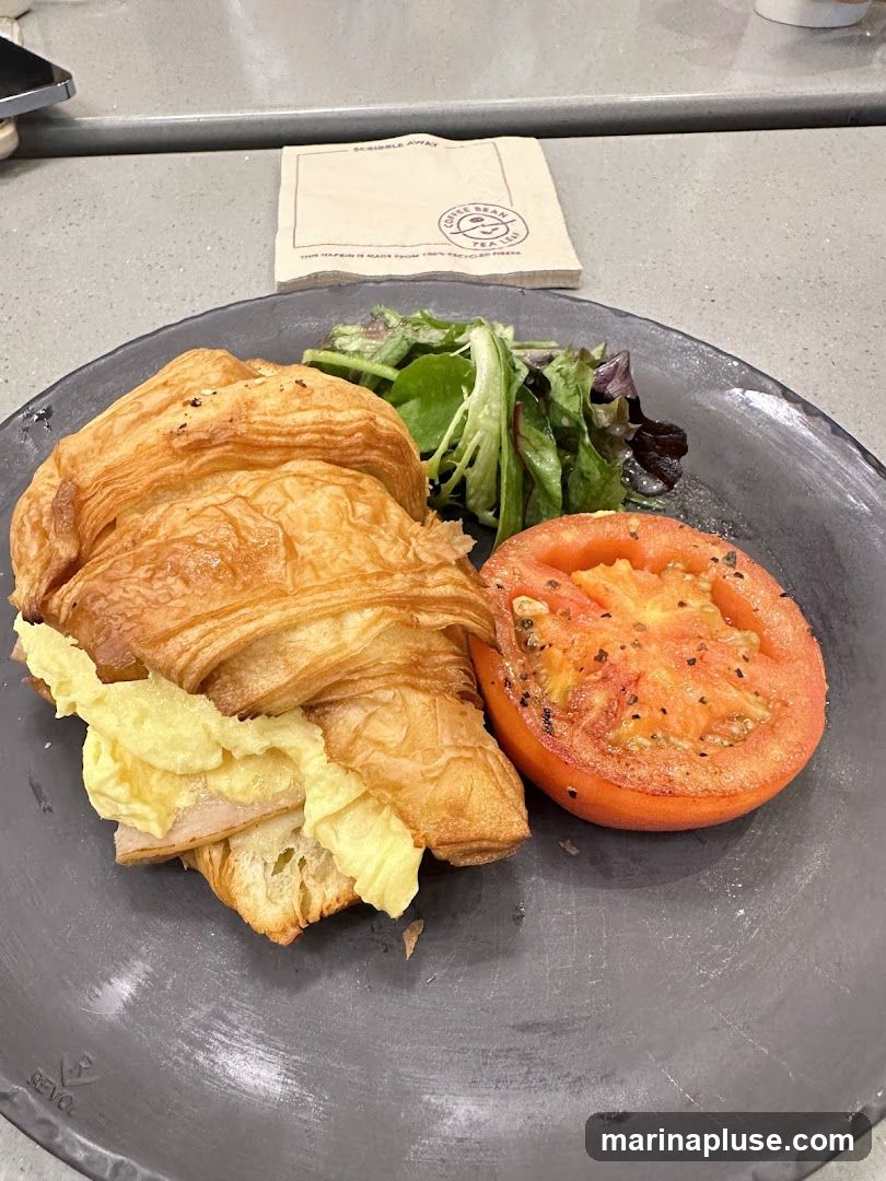 A close-up shot of a croissant sandwich filled with scrambled eggs and ham, served with a slice of grilled tomato and a small side salad on a dark plate. A napkin with the Coffee Bean & Tea Leaf logo is visible in the background.