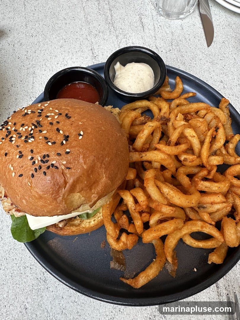 A close-up shot of a burger served with curly fries, ketchup, and a dipping sauce. The burger has a sesame seed bun and appears to have lettuce and cheese inside. The curly fries are golden brown and seasoned.