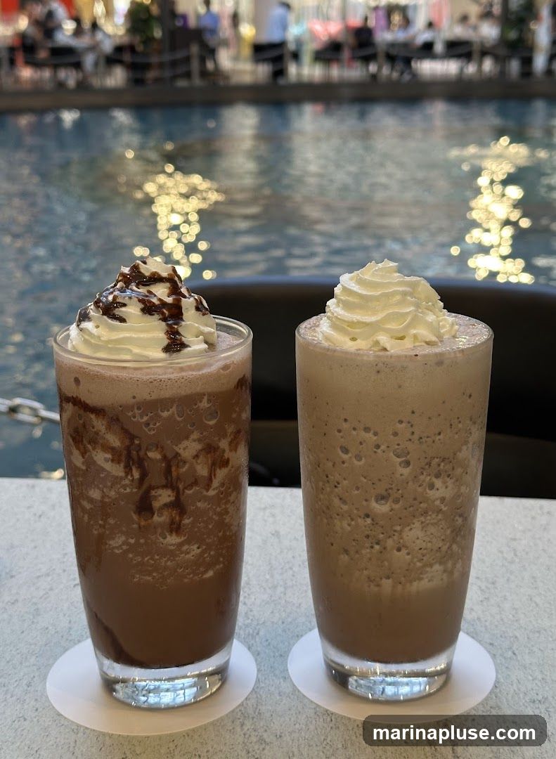Two tall glasses of chocolate frappes topped with whipped cream sit on a table. One is drizzled with chocolate sauce. In the background, a body of water reflects lights, and blurred figures are visible seated at tables.
