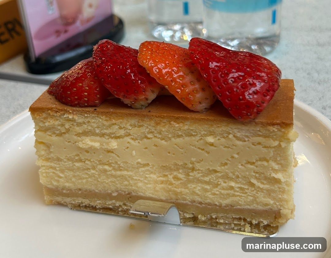 A slice of cheesecake topped with fresh strawberries sits on a white plate. The cheesecake has multiple layers of creamy filling and a golden-brown cake base. In the background, blurred bottles of water and a menu stand are visible.