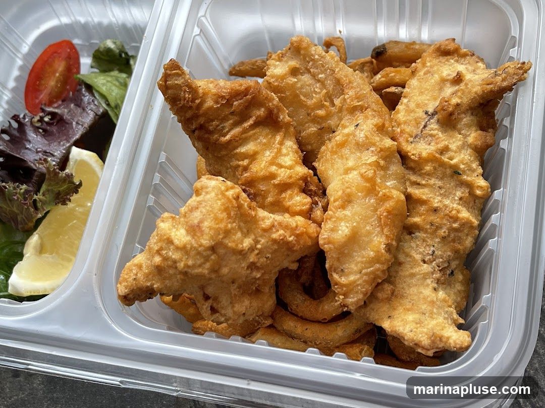 A close-up shot of a takeout container filled with fried fish pieces and curly fries. A small side salad with lettuce, a slice of tomato, and a lemon wedge is visible on the left.