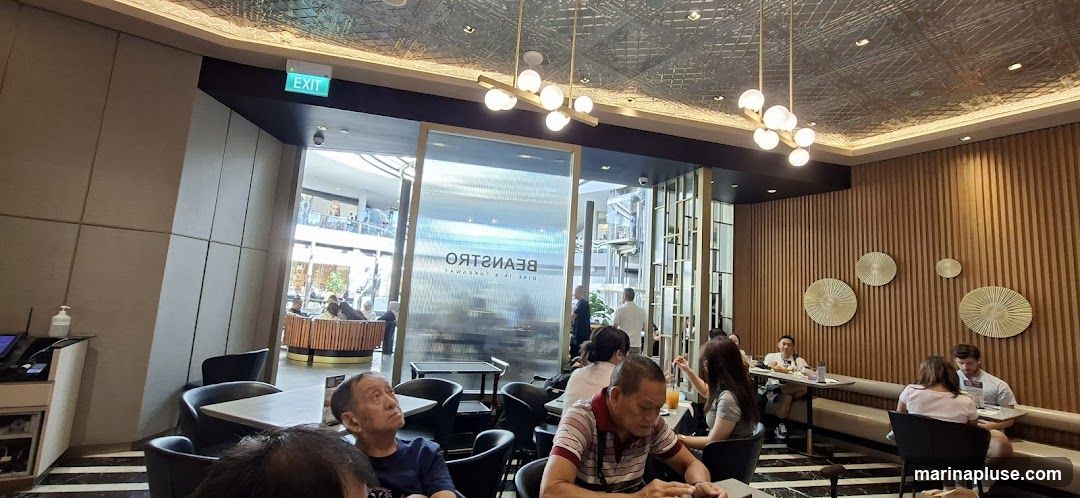The image shows the interior of a modern cafe with a sleek design. The walls are decorated with vertical wooden slats and circular metallic art pieces. The ceiling has an intricate patterned design and is illuminated by contemporary pendant lights. Several tables with chairs are arranged throughout the space, and patrons are seen dining and conversing. A large glass partition with the reversed text "BEANSTRTO" and "DINE IN & TAKEAWAY" is visible, suggesting the name of the establishment. An "EXIT" sign is also visible above a doorway.