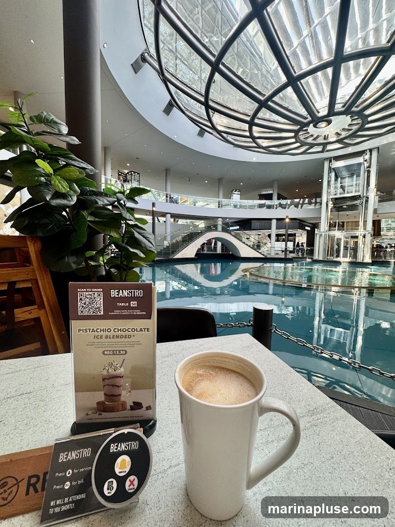 A white mug of coffee sits on a table in front of a menu for Beanstro. The menu features a Pistachio Chocolate Ice Blended drink. In the background, a large indoor atrium with a water feature and multiple levels of shops is visible.