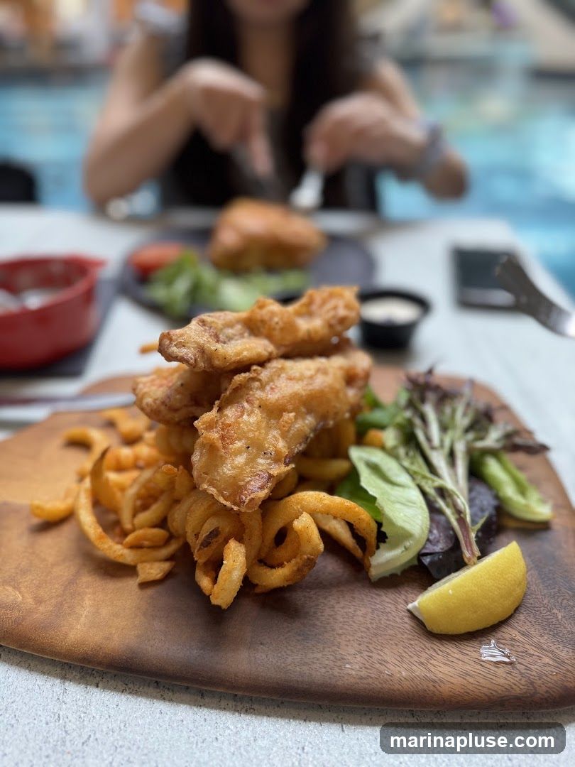 A close-up shot of a plate of fish and chips served on a wooden board. The dish includes several pieces of battered fish, curly fries, a small side salad with lettuce and tomato, and a lemon wedge. A person is visible in the blurred background, using cutlery to eat.