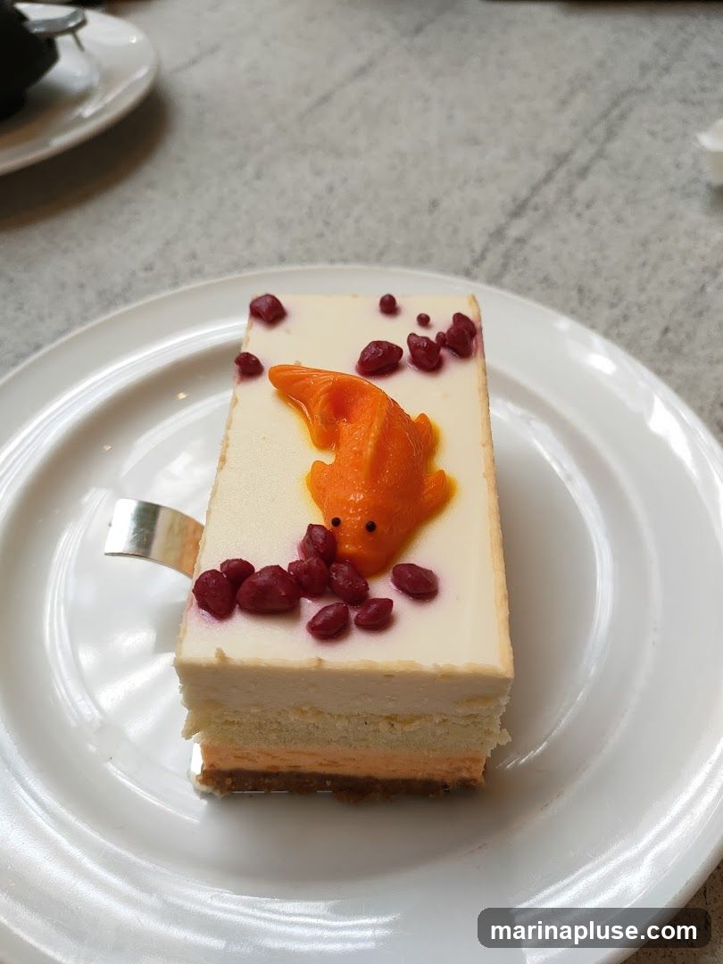 A rectangular slice of layered cake is presented on a white plate. The cake has a white frosting on top, decorated with a bright orange koi fish made of icing and several small, dark red berries. The cake itself appears to have multiple layers of different colors, including white, light orange, and a darker orange or brown base. A silver utensil is partially visible on the left side of the plate. The background is a textured, light-colored surface, possibly a table.