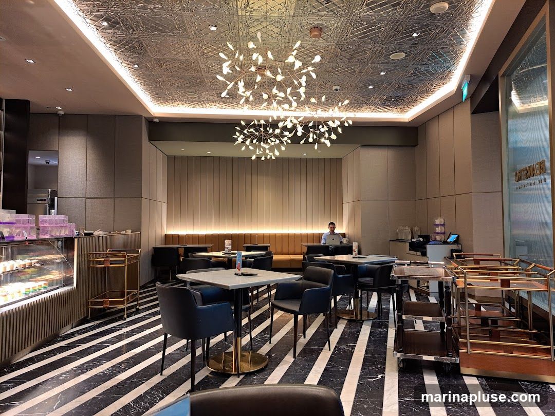 A modern cafe interior with a striking black and white striped marble floor. Tables with dark chairs are arranged throughout the space, some with a view of a display case filled with colorful desserts. A man is seated at a table in the background, working on a laptop. The ceiling features an ornate metallic design and a contemporary chandelier. Service carts are visible near the entrance.