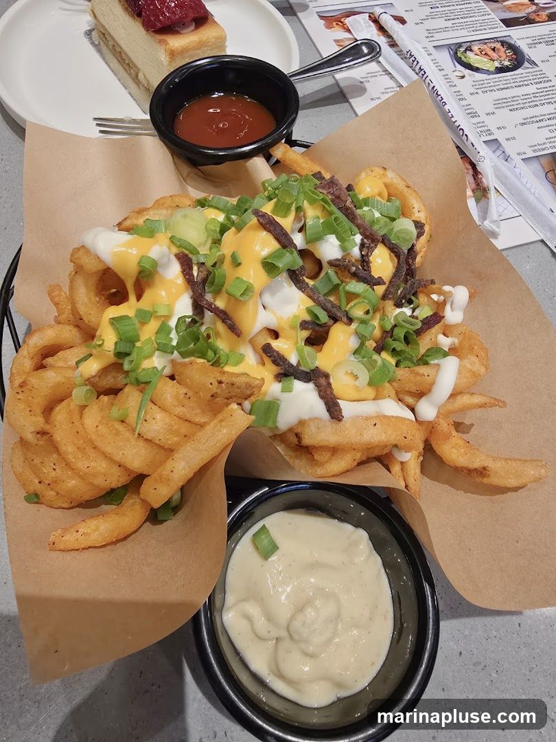 A basket of curly fries topped with cheese sauce, sour cream, bacon bits, and green onions is served with a side of dipping sauce. A slice of cheesecake with strawberry topping sits on a white plate in the background. A menu is visible on the right side of the image.