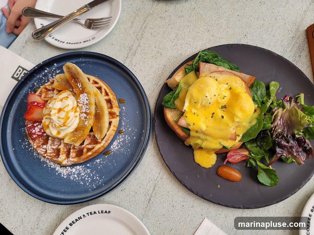 A close-up view of two brunch dishes on a table. On the left, a waffle topped with whipped cream, sliced bananas, strawberries, and caramel sauce. On the right, Eggs Benedict served with ham, spinach, and a side salad. A plate with cutlery is visible in the background, and a small plate with the logo "THE COFFEE BEAN & TEA LEAF" is in the foreground.