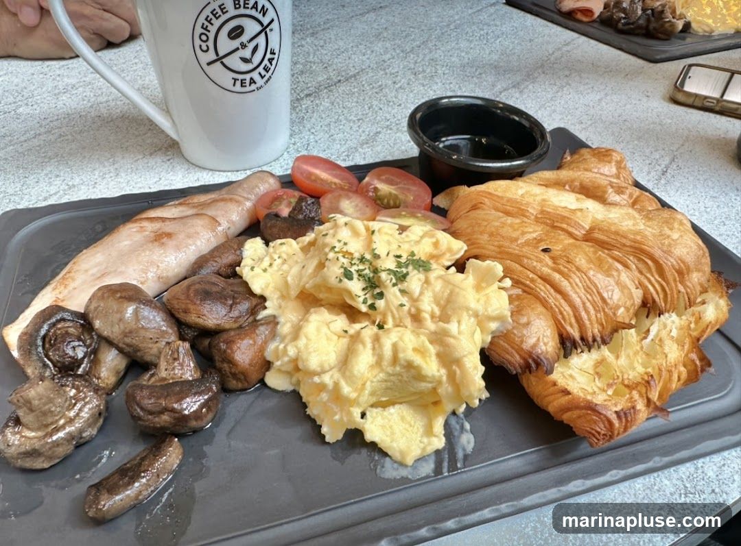A breakfast platter with scrambled eggs, a croissant, sausage, mushrooms, and cherry tomatoes, served with a mug of coffee.