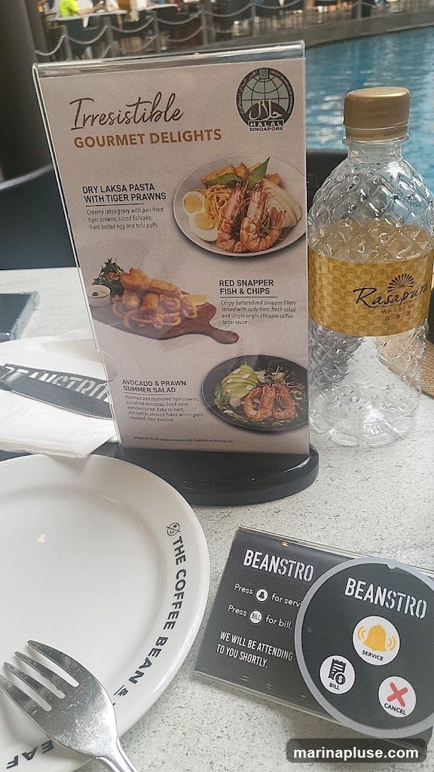 A menu displays gourmet delights including Dry Laksa Pasta with Tiger Prawns, Red Snapper Fish & Chips, and Avocado & Prawn Summer Salad. A call button for Beanstro is also visible, with options for service, bill, and cancel. A bottle of Rasapuri water and a plate from The Coffee Bean are on the table.