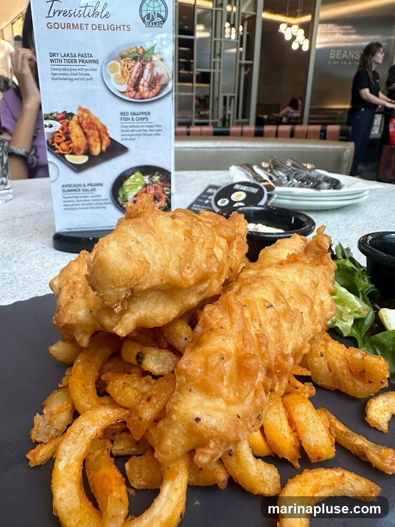 A close-up of a plate of fish and chips with curly fries, next to a menu board displaying various gourmet delights. The menu features Dry Laksa Pasta with Tiger Prawns, Red Snapper Fish & Chips, and Avocado & Prawn Summer Salad.