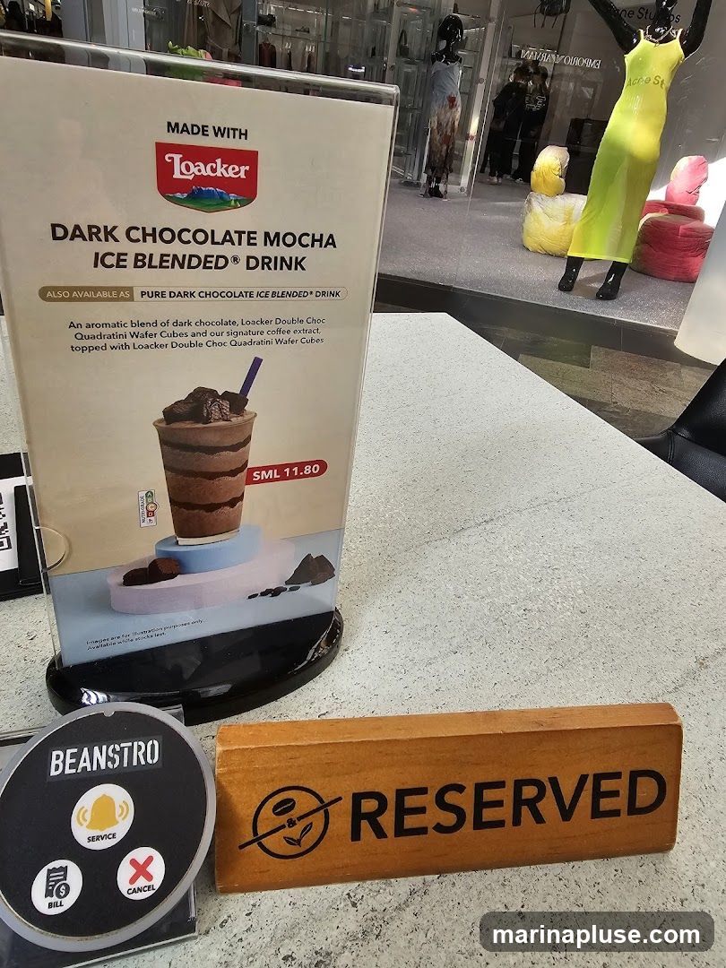A sign advertising a Loacker Dark Chocolate Mocha Ice Blended Drink, also available as a Pure Dark Chocolate Ice Blended Drink. The description mentions an aromatic blend of dark chocolate, Loacker Double Choc Quadratini Wafer Cubes, and signature coffee extract. A small sign indicates "BEANSTRO" with options for "SERVICE", "BILL", and "CANCEL". A wooden "RESERVED" sign is also visible. In the background, a store window displays mannequins and merchandise.