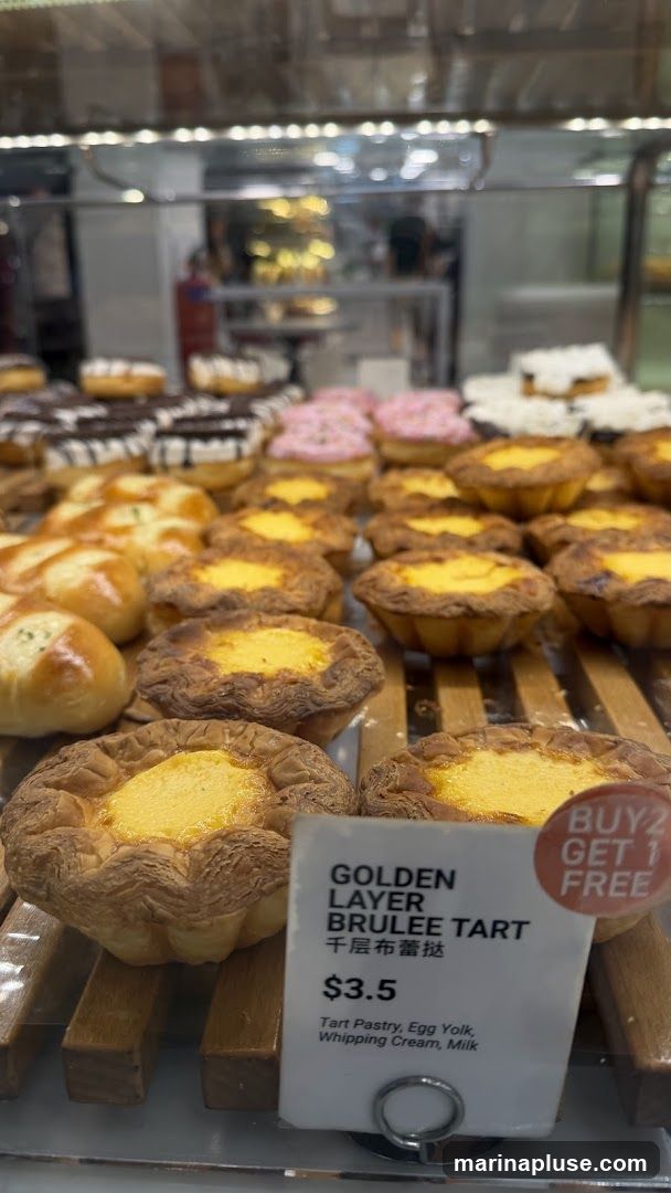 A close-up shot of a display case filled with various pastries, including donuts and tarts. In the foreground, several golden-brown tarts with a creamy yellow filling are prominently featured. A price tag indicates that a "Golden Layer Brulee Tart" costs $3.5 and is made with tart pastry, egg yolk, whipping cream, and milk. A promotional sticker suggests a "Buy 2 Get 1 Free" offer.