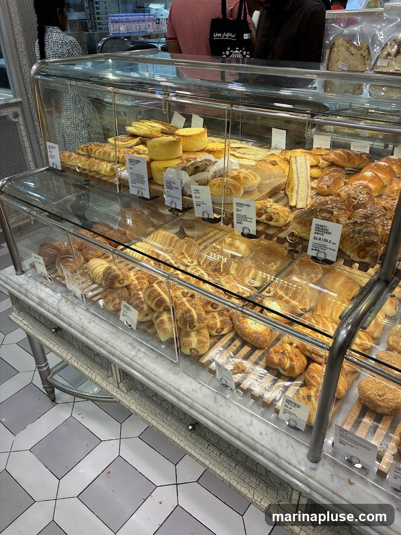 A display case filled with various baked goods, including bread, pastries, and sandwiches. The items are arranged on wooden trays and shelves behind glass. Some items have small labels with names and prices.