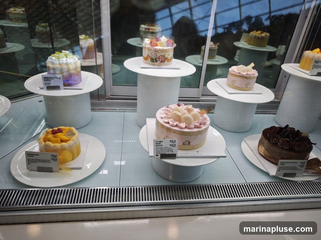 A display case filled with various cakes and pastries. In the foreground, there are several individual cakes on white plates. One cake is yellow with mango and red berries. Another is pink with lychee and floral decorations. A dark chocolate cake is also visible. In the background, more cakes are displayed on white stands, some with fruit toppings. The display is behind glass, reflecting the environment outside.