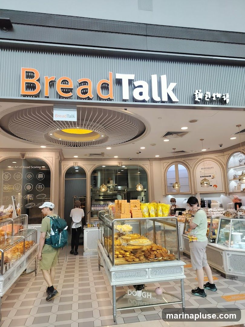 The interior of a BreadTalk bakery with displays of bread and pastries. Customers are browsing the selection. The shop features a modern design with clean lines and bright lighting.