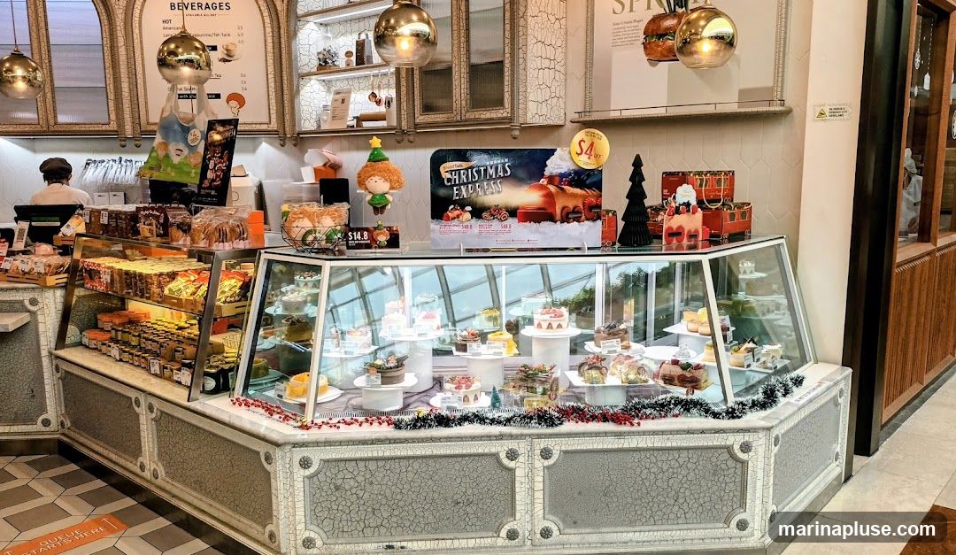 A bakery display case filled with various cakes and pastries, decorated for Christmas. A promotional poster for a 'Christmas Express' cake is visible, along with festive figurines and gift boxes. Packaged goods and jars are displayed on shelves behind the main display.