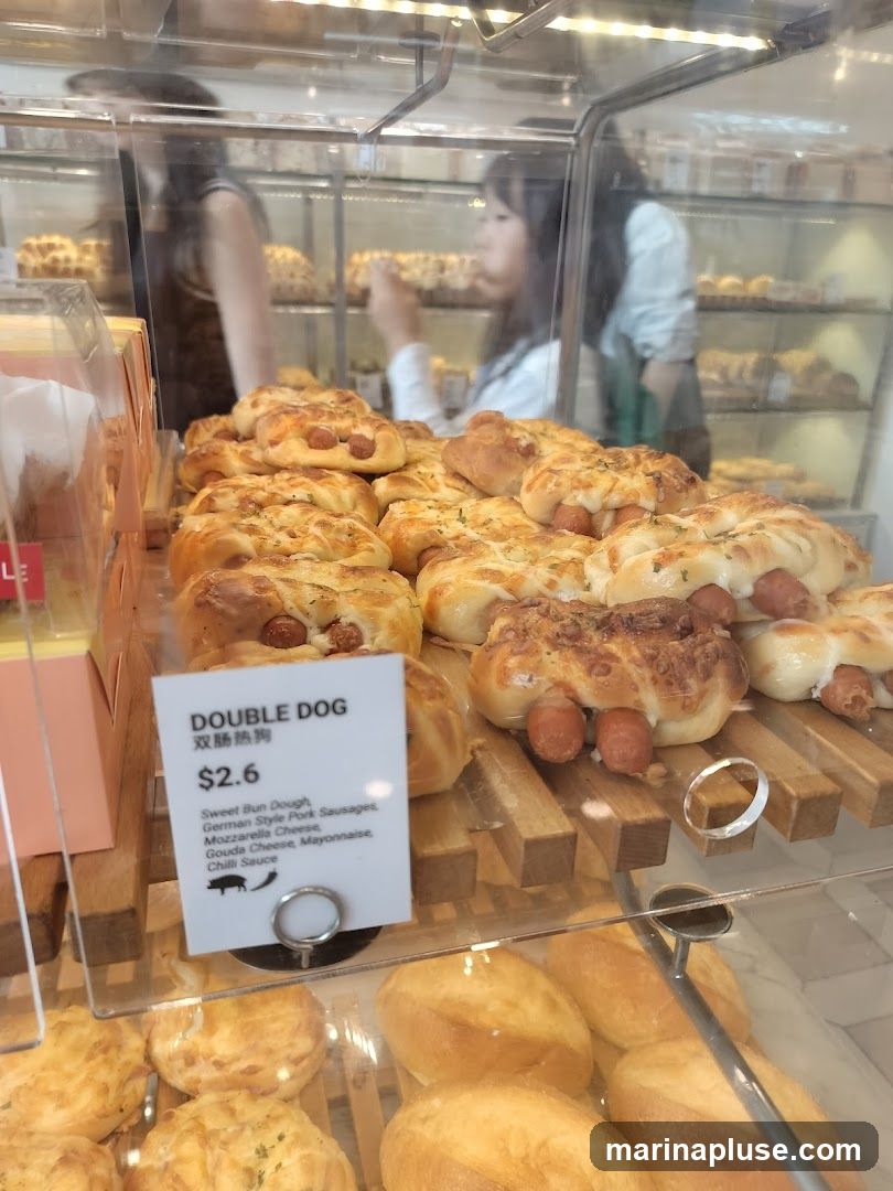 A display case filled with various baked goods, prominently featuring a pastry labeled "DOUBLE DOG" priced at $2.6. The pastry appears to be a sweet bun dough filled with German-style pork sausages and topped with cheese, mayonnaise, and chili sauce. Other pastries and bread are visible in the background and foreground.