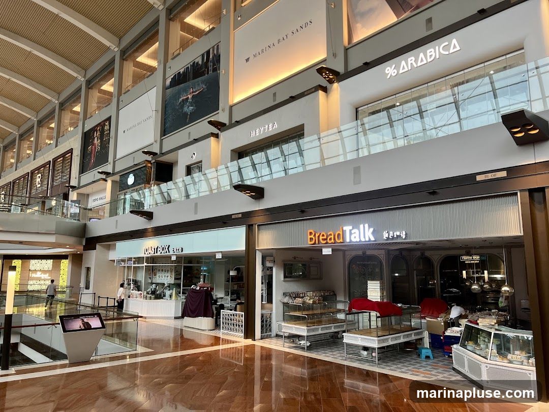 An interior view of a modern shopping mall with multiple storefronts visible on the lower level. Storefronts include "Toast Box", "BreadTalk", and "% Arabica". Large advertisements and signage for "Marina Bay Sands" and "Heytea" are displayed on the upper levels. The mall features a polished floor and a multi-level design with glass railings.