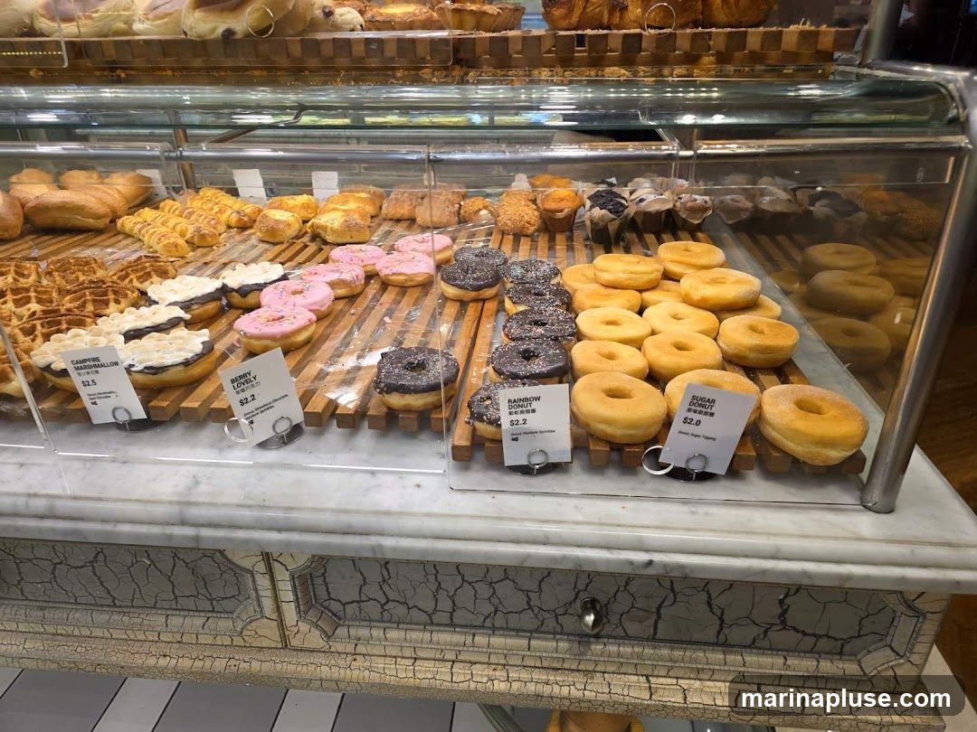 A display case filled with various baked goods, including donuts, pastries, and bread. The donuts are arranged in rows, with some topped with chocolate and sprinkles, and others plain or sugared. There are also waffles and pastries with marshmallow topping. Several small signs indicate the names and prices of some items.