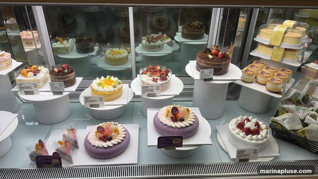 A refrigerated display case filled with various cakes, pastries, and desserts. There are whole cakes, slices of cake, and individual puddings. Some cakes are decorated with fruit and cream. Labels with prices are visible for some items.