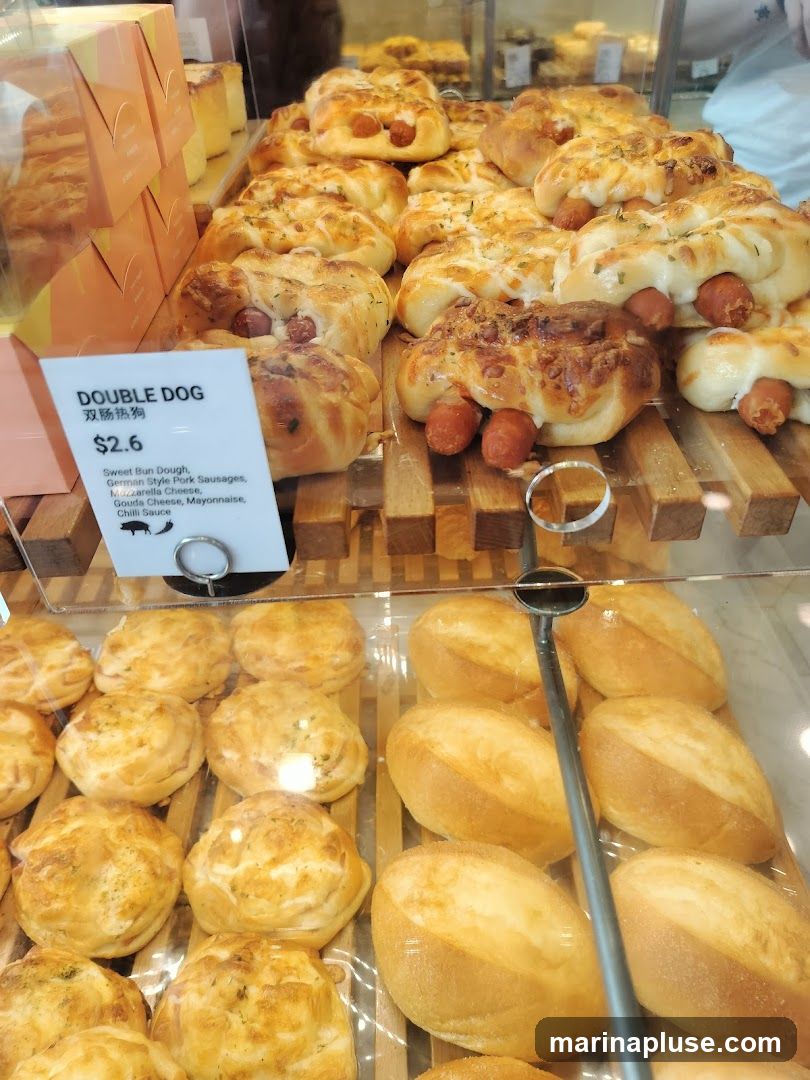 A close-up view of a bakery display case filled with various baked goods. In the foreground, there are rows of bread rolls and savory buns. Prominently featured are several "Double Dog" buns, which are sweet buns filled with sausages and topped with cheese and herbs. A label indicates the price and ingredients for the "Double Dog" bun.