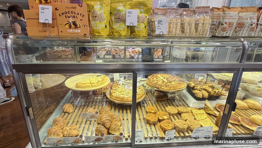 A display case filled with various baked goods, including pizzas, quiches, croissants, and bread. Packaged snacks like chocolate swirls and lemon wafers are visible on top.