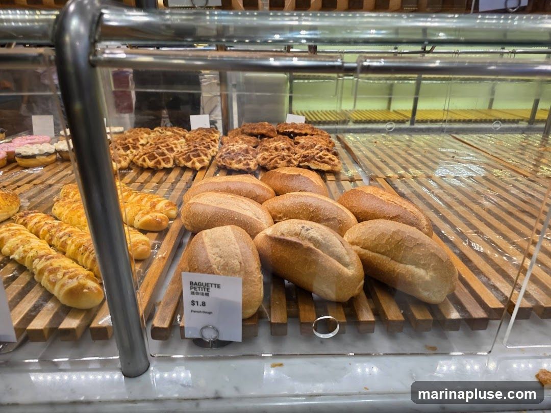 A close-up view of a bakery display case filled with various baked goods. In the foreground, several loaves of bread are arranged on wooden shelves. A small white sign indicates "BAGUETTE PETITE" for $1.8, described as "French Dough." Behind these loaves, there are rows of golden-brown pastries, some resembling waffles and others with a flaky texture. Further back, donuts topped with pink and white frosting are visible. The display case has a glass front and metal supports.