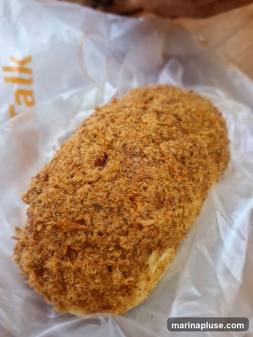 A close-up shot of a bread roll covered in pork floss. The bread is oval-shaped and appears to be soft. The pork floss is finely shredded and has an orange-brown color. The bread is wrapped in a white plastic bag with orange lettering that is partially visible, reading "alk".