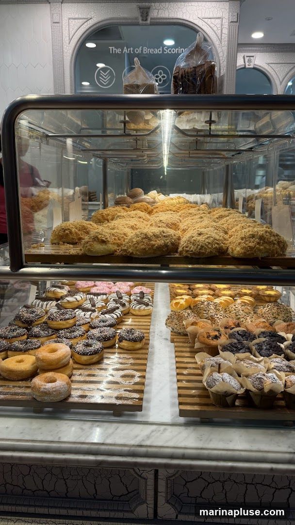 A display case filled with various baked goods, including donuts, muffins, and bread. The top shelf features bread rolls covered in a shredded topping. The middle shelf has an assortment of donuts, some plain, some with chocolate frosting and sprinkles, and others with chocolate drizzle and a candy bar piece. The bottom shelf displays muffins and other pastries. Above the display, a sign reads "The Art of Bread Scoring.".