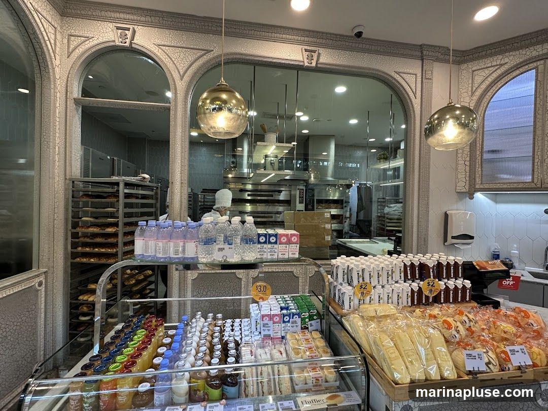 A view inside a bakery, showcasing a display of various pastries, bread, bottled drinks, and cartons of milk. In the background, bakers can be seen working in a kitchen area with ovens and racks of baked goods. The decor features ornate archways and hanging pendant lights.