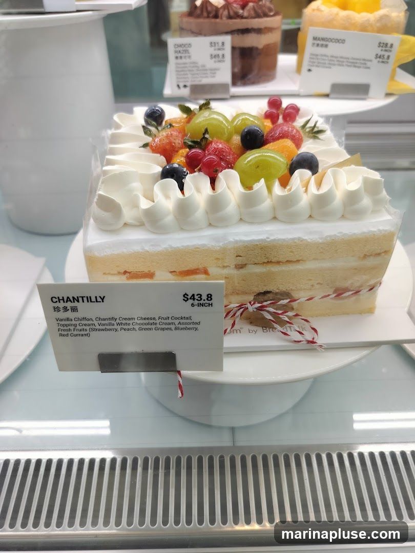 A Chantilly cake with assorted fresh fruits on top, displayed in a refrigerated case. The cake has layers of vanilla chiffon, chantilly cream cheese, and fruit cocktail, topped with cream and white chocolate cream. The fruits include strawberries, peaches, green grapes, blueberries, and red currants. A label indicates the price is $43.8 for a 6-inch cake. Other cakes, including a chocolate hazelnut cake and a mango coco cake, are visible in the background.