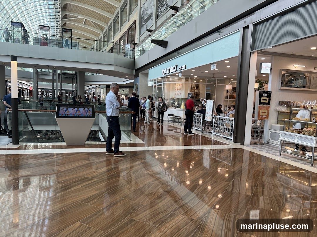A wide shot of a modern indoor shopping mall with a polished wooden floor reflecting the lights. In the mid-ground, several shops are visible, including one with a prominent sign that reads "TOAST BOX". People are seen walking and queuing at the shops. An information kiosk with a screen is also present. The architecture features a high, vaulted ceiling with a glass roof and multiple levels of walkways and shops.