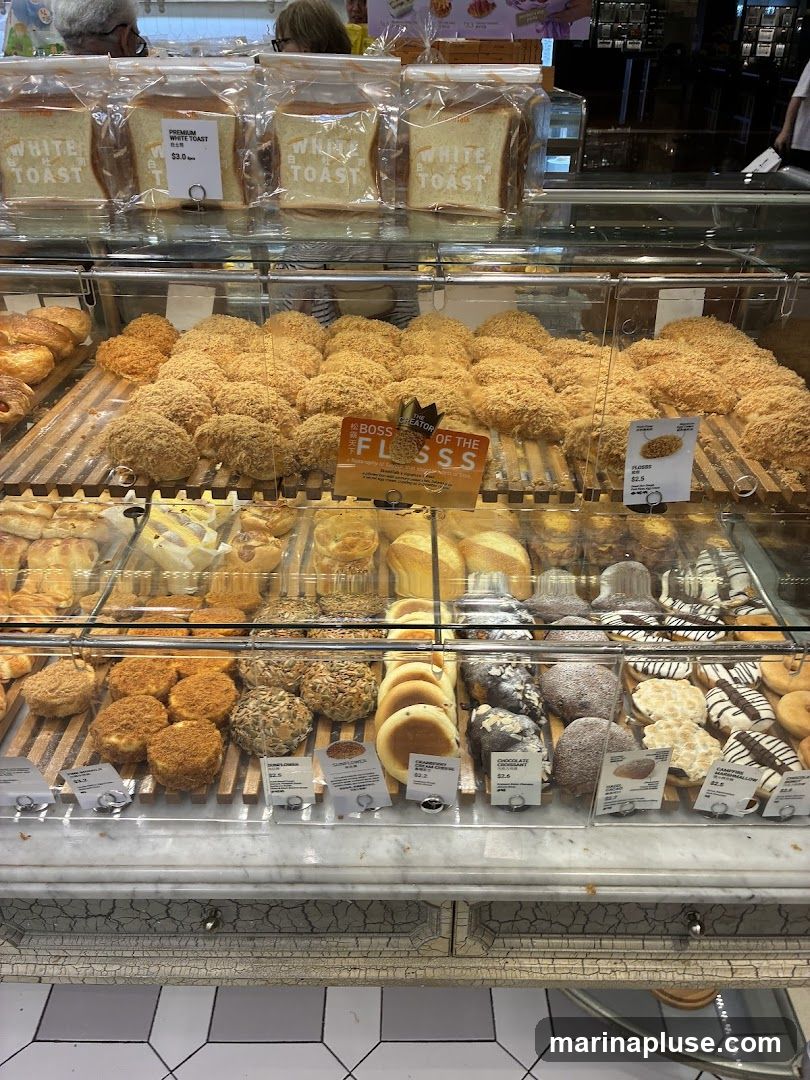 A display case filled with various baked goods, including loaves of white toast, fluffy buns topped with shredded meat, round pastries, and donuts. A sign reads "BOSS OF THE FLOSSS".