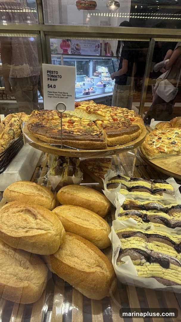 A close-up view of a bakery display case filled with various baked goods. In the foreground, several loaves of bread are arranged. Behind them, a large pizza is displayed on a wooden stand, with a sign indicating it is Pesto Tomato flavor for $4.0. The sign also lists the ingredients: Pizza Bread, Pizza Sauce, Cheese, Tomato, Pesto Sauce, Virgin Olive Oil, Basil Leaf. Further back, more pastries and cakes are visible in refrigerated display cases, along with customers browsing.