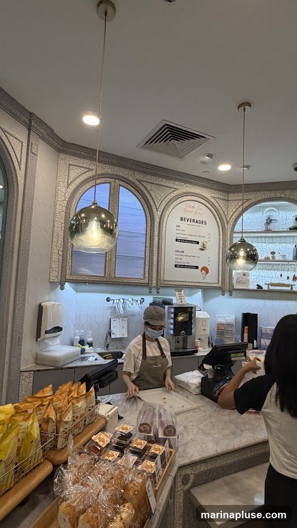 A view of a bakery counter with various baked goods on display. A staff member wearing an apron and mask is visible behind the counter, preparing an order. Above the counter, a menu board lists beverages. The interior features decorative arched windows and pendant lights.
