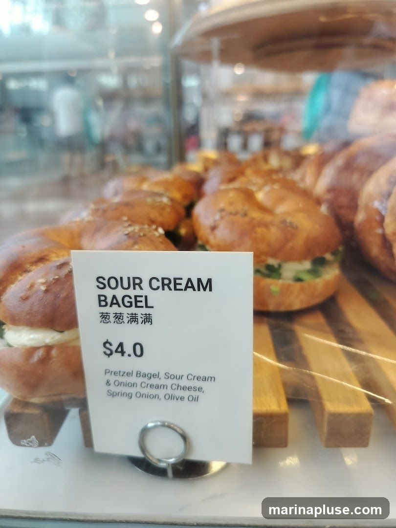 A close-up of a sour cream bagel with a price tag in front of it. The bagel appears to be a pretzel bagel topped with sesame seeds and filled with sour cream and green onions. The price tag indicates it costs $4.0 and lists the ingredients as Pretzel Bagel, Sour Cream & Onion Cream Cheese, Spring Onion, Olive Oil. In the background, other baked goods are visible in a display case.