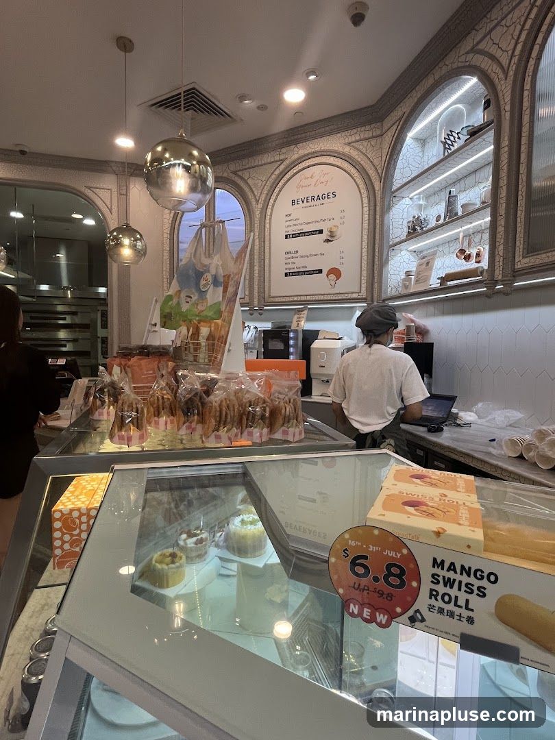 An interior view of a bakery with display cases filled with pastries and cakes. A beverage menu is visible on the wall, listing hot and chilled drinks. A staff member is seen working behind the counter. A promotional sign for a Mango Swiss Roll is displayed.