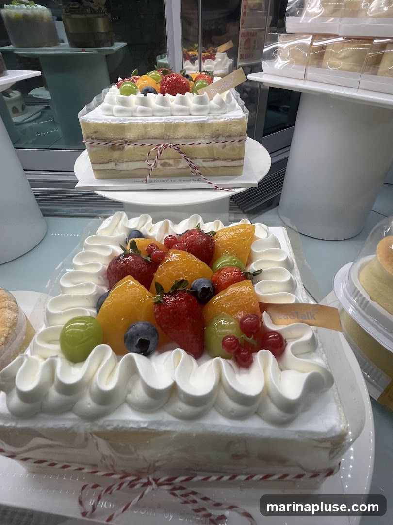 A display of two fruit-topped cakes in a bakery case. The cake in the foreground is square with white frosting piped around the edges and topped with strawberries, blueberries, grapes, peaches, and red currants. The cake behind it is also square and decorated similarly. Other cakes and pastries are visible in the background. A small tag on the cake in the foreground reads "BreadTalk" and "the icing room".